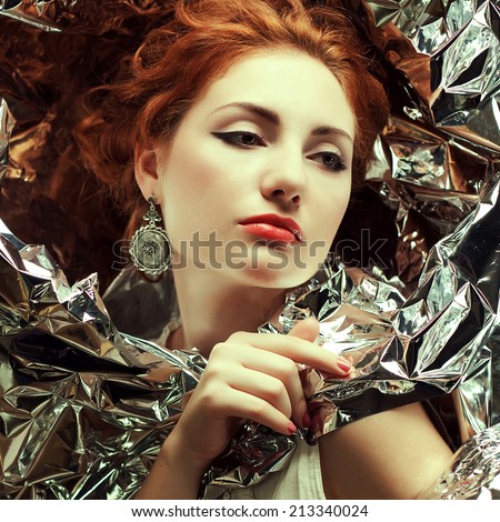 Arty portrait of sad fashionable queen-like red-haired (ginger) model with silver foil cape. Silver vintage earrings. Perfect make-up. Retro style. Close up. Studio shot