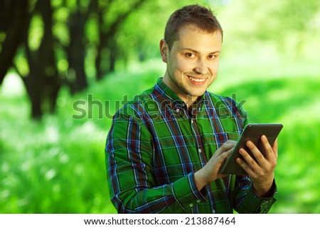 Gadget user concept. Portrait of funny hipster guy in green casual shirt reading e-book in the park. White shiny smile. Bristle on face with healthy skin. Copy-space. Hipster style. Outdoor shot