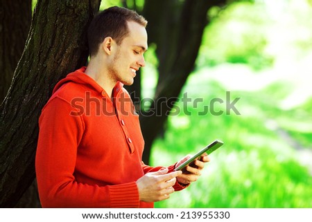 Gadget user concept. Portrait of happy young guy in orange sweater reading e-book in the park. White shiny smile. Bristle on face with healthy skin. Copy-space. Hipster style. Outdoor shot