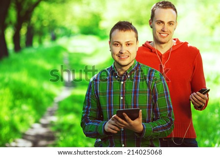 Gadget user concept. Funny hipster guys in trendy casual clothing reading e-book, listening music in park. White shiny smile. Bristle on face with healthy skin. Copy-space. Hipster style. Outdoor shot