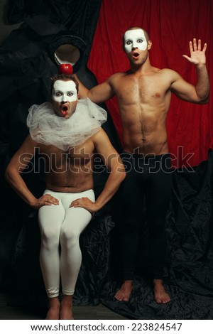 Hocus pocus concept. Two mime artists, clowns with white masks on faces performing focus with hat and red apple over red cloth & black background. Magician and victim of trick. Wow faces. Studio shot