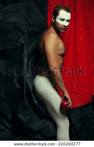 Backstage concept. Arty portrait of circus performer in white tights holding red apple, posing over black and red cloth. White mask on face. Muscular body and perfect tan. Halloween party. Studio shot