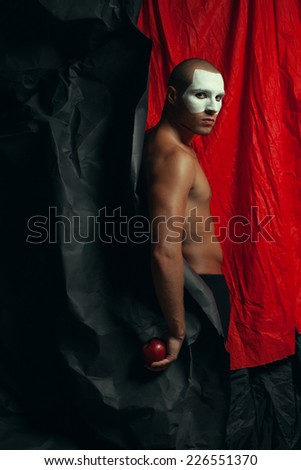 Backstage concept. Arty portrait of circus performer in black tights holding red apple, posing over black and red cloth. White mask on face. Muscular body and perfect tan. Halloween party. Studio shot