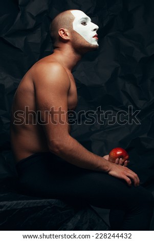 Backstage concept. Arty portrait of circus performer in black tights holding red apple, posing over black cloth. White mask on face. Muscular body and perfect tan. Halloween party. Studio shot