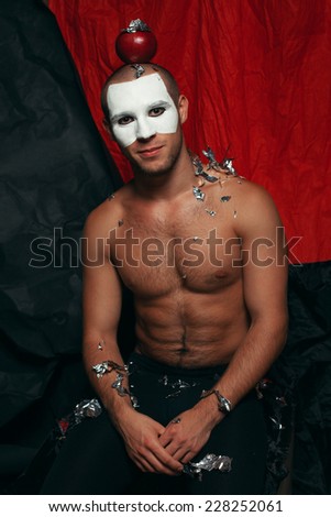 Backstage concept. Arty portrait of circus performer in black tights with red apple on head and sequins on body. White mask on face. Muscular body and perfect tan. Halloween party