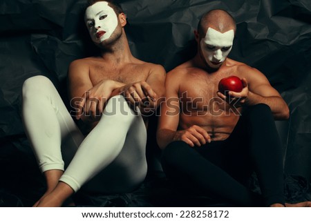 Backstage concept. Arty portrait of two circus performers in tights holding red apple, posing over black cloth. White masks on faces. Muscular bodies and perfect tan. Halloween party