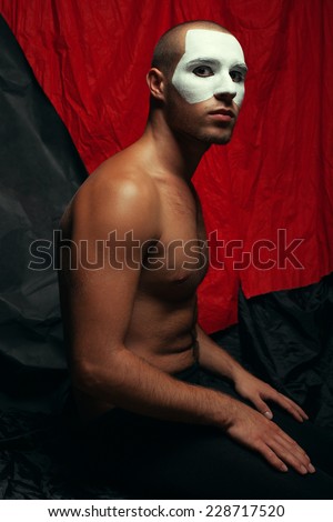 Backstage concept. Arty portrait of circus performer in black tights posing over black and red cloth. White mask on face. Muscular body and perfect tan. Halloween party. Studio shot