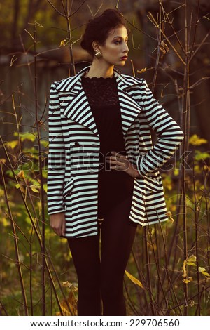 Emotive portrait of Hollywood diva, film star walking in the park. Evening time. Sunny spring weather. Young woman in luxurious trendy striped jacket. Golden earrings. Outdoor shot
