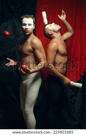 Freak circus concept. Two muscular mime artists, clowns with white masks on faces juggling and posing over red cloth & black background. Vintage style. Studio shot