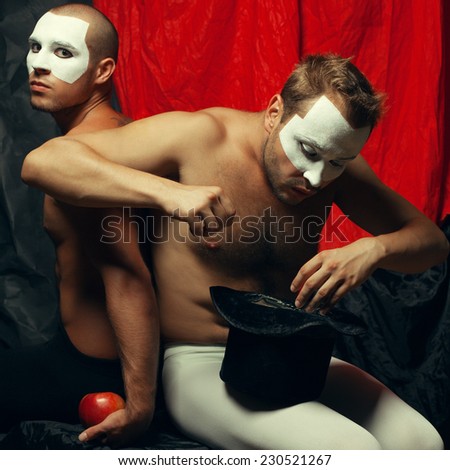 Hocus pocus concept. Two mime artists, clowns with white masks on faces performing focus with hat and red apple over red cloth & black background. Studio shot