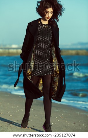 High fashion concept. Emotive portrait of beautiful brunette with long curly hair wearing black coat and little black dress & walking along beach. Windy weather. Italian luxurious style. Outdoor shot