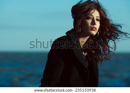 Emotive portrait of beautiful brunette with long curly hair in black coat. Luxurious golden accessories: earrings, necklace. Perfect make-up. Vogue style. Copy-space. Windy weather. Outdoor shot