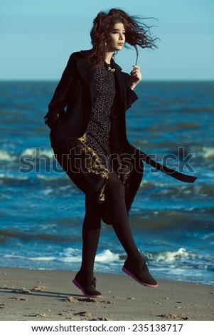 High fashion concept. Emotive portrait of beautiful brunette with long curly hair wearing black coat and little black dress & walking along beach. Windy weather. Italian luxurious style. Outdoor shot
