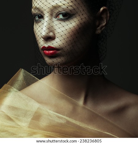 Art fashion concept. Portrait of sensual beautiful brunette looking at camera through black veil and posing over dark gray background. Golden eyebrows, red wet lips. Close up. Vogue style. Studio shot