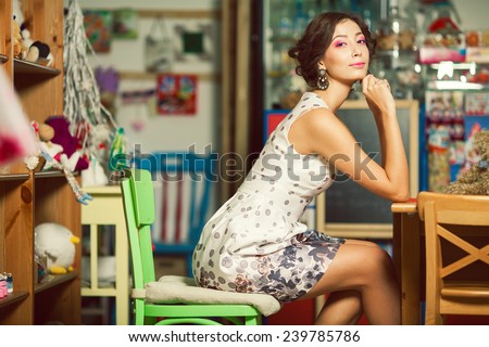 Candy girl concept. Emotive portrait of happy beautiful doll like woman with perfect make-up, hairdo, diamond earrings sitting in cafe with vintage decoration. Indoor shot