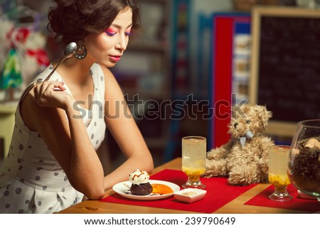 Guilty pleasure concept. Portrait of doll like brunette girl with retro hairdo in white trendy vintage polka-dot sleeveless dress eating cake, drinking tea in cafe. Close up. Copy-space. Indoor shot