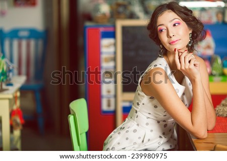 Candy girl concept. Portrait of happy beautiful doll like woman with perfect make-up, hairdo, diamond earrings sitting in cafe with toys and vintage decoration. Indoor shot