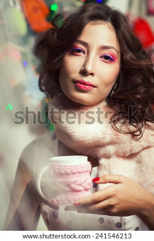 Winter holidays celebration concept. Portrait of doll like brunette in retro polka-dot sleeveless dress drinking tea or coffee in restaurant, cafe. Pink fur scarf on her neck. Close up. Indoor shot