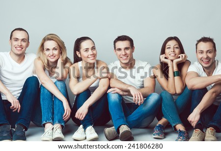 Happy together concept. Group portrait of healthy boys and girls in white t-shirts, sleeveless shirts and blue jeans sitting and posing over gray background. Copy-space. Urban style. Studio shot