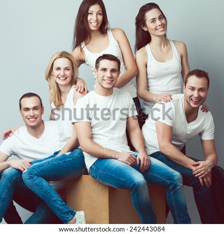 Happy together concept. Group portrait of healthy boys & girls in white t-shirts, sleeveless shirts and blue jeans standing, sitting, posing over gray background. Urban street style. Studio shot
