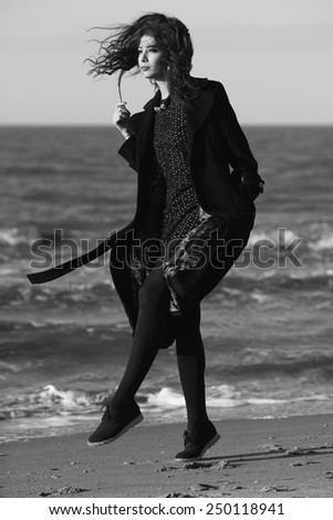 High fashion concept. Portrait of beautiful brunette with long curly hair wearing black coat and little black dress & feeling happy on the beach. Sunny weather. Italian luxurious style. Outdoor shot
