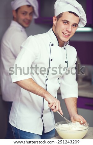 Dessert cooking concept. Portrait of a smiling male chef with his coworker cooking food and standing in the modern kitchen of restaurant. Indoor shot