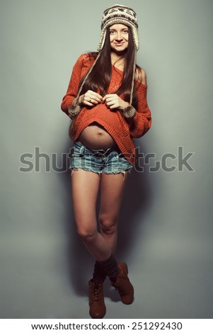 Stylish pregnancy concept. Portrait of happy pregnant woman with hands on stomach posing in trendy clothing and leather shoes over gray background. Casual style. Studio shot