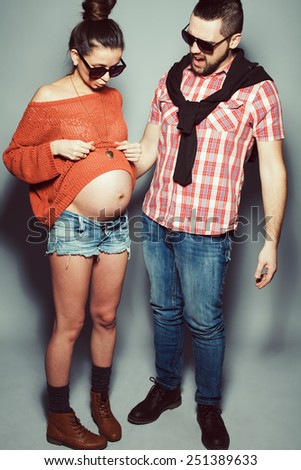Stylish pregnancy & family concept: portrait of funny couple of hipsters (husband and wife) in trendy casual clothing, eyewear posing over gray background. Urban street style. Studio shot