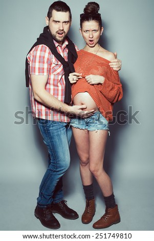 Stylish pregnancy & family concept: portrait of disturbed couple of hipsters (husband and wife) in trendy casual clothing, eyewear posing over gray background. Urban street style. Studio shot
