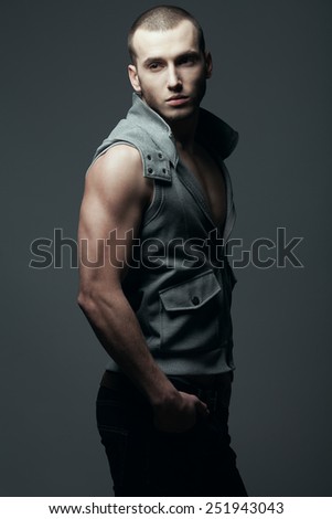 Portrait of handsome brutal young man with short hair and bristle on face wearing sleeveless jacket, jeans and posing over dark gray background. Hipster style. Studio shot