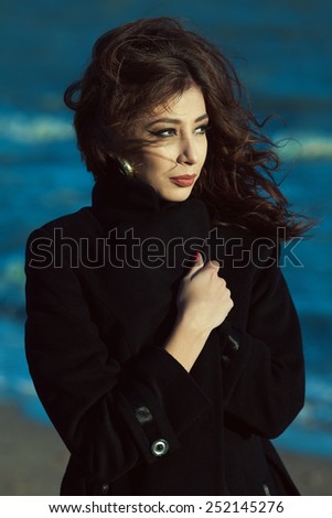 Emotive portrait of beautiful brunette with long curly hair hiding in black coat and walking at the seaside. Luxurious golden earrings. Street and vogue style. Windy weather. Outdoor shot