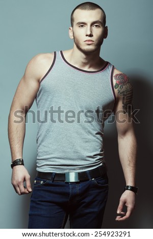 Portrait of tattooed brutal young man with short hair and bristle on face wearing sleeveless shirt, blue jeans and posing over gray background. Hipster style. Studio shot