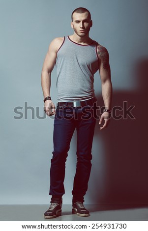 Full length portrait of tattooed brutal young man with short hair, bristle on face wearing sleeveless shirt, blue jeans, sneakers, posing over gray background. Hipster style. Studio shot