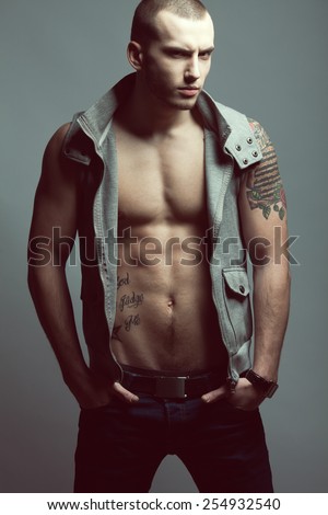 Portrait of tattooed brutal young man with short hair and bristle on face wearing sleeveless jacket, blue jeans and posing over gray background. Hipster style. Studio shot