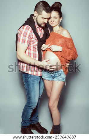 Stylish pregnancy & family concept: portrait of loving couple of hipsters (husband and wife) in trendy casual clothing posing over gray background. Urban street style. Studio shot