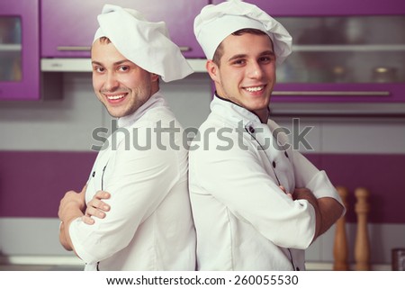 Portrait of two funny working men in cook uniform posing in modern kitchen. Indoor shot