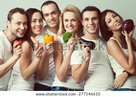 Happy veggies concept. Group portrait of healthy boys and girls in white t-shirts, sleeveless shirts and blue jeans standing with vegetables & posing over gray background. Urban style. Studio shot
