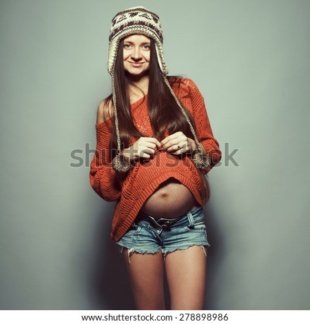 Stylish pregnancy concept. Portrait of happy hipster mommy with hands on stomach posing in trendy clothing over gray background. Casual style. Studio shot