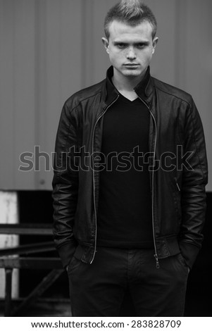 Bad boy concept. Portrait of brutal young man with short hair wearing black jacket, jeans and posing over urban background. Hipster style. Outdoor shot