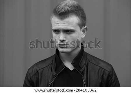 Stylish bully concept. Portrait of brutal young man with short wet hair wearing black jacket and posing over urban background. Hipster style. Close up. Black and white outdoor shot
