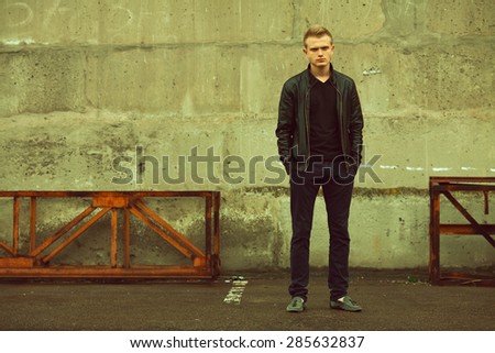 Bad boy concept. Portrait of brutal young man with short hair wearing black jacket, jeans and posing over urban background. Hands in pockets. Hipster style. Copy-space. Outdoor shot