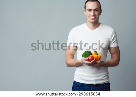 Raw, living food concept. Portrait of happy young man wearing white t-shirt, blue jeans, holding vegetables in hands over gray background. Casual clothing. Muscular body. Copy-space. Studio shot