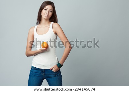 Raw, living food concept. Portrait of happy young woman wearing white sleeveless shirt, jeans, holding orange over gray background. Casual clothing. Fit body. Natural make-up. Copy-space. Studio shot