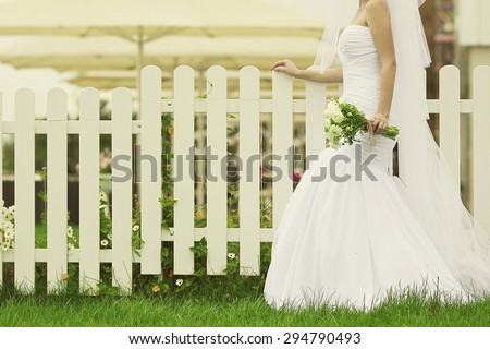Wedding trends concept. Bride\'s hands with bouquet of beige flowers over white wooden fence and green lawn. Trendy dress with vapory veil. Country vintage style. Copy-space. Outdoor shot