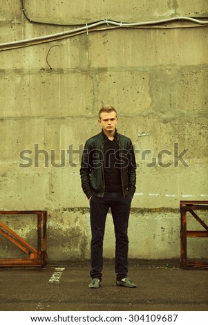 Bad boy concept. Full length portrait of brutal young man with short hair wearing black jacket, jeans and posing over urban background. Hands in pockets. Hipster style. Copy-space. Outdoor shot