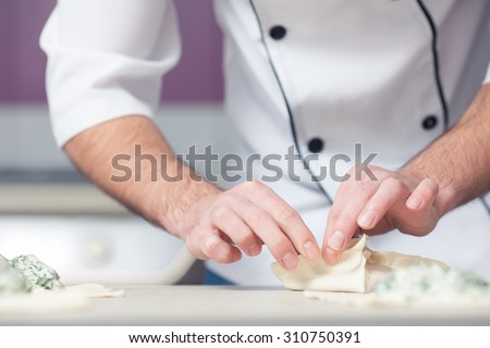 Vegetarian bakery concept. Chef cook in uniform putting filler of spinach and soft feta cheese on slice of puff pastry and making french chausson. Close up. Indoor shot