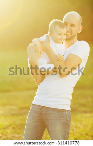 Happy family concept. Portrait of playing father and little son in white casual t-shirts. Toddler in white socks and diaper. Rays of light at summer sunrise. Outdoor shot