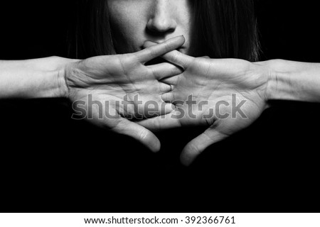 Closeup portrait of young woman hiding mouth by hands, showing speak no evil concept, isolated on black background. Human emotion, expression, rights, communication. Copy-space. Monochrome studio shot