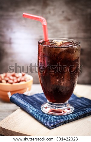 glass of cola with ice and salt roasted peanuts have lighting beside on wood table