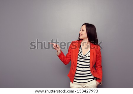 cheerful young woman in red jacket pointing at empty copyspace over grey background
